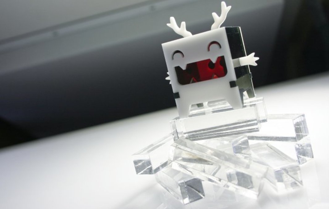 A photo of a boxy, acrylic designer toy with a toothy grin and antlers standing on a pile of translucent colorless bits of acrylic