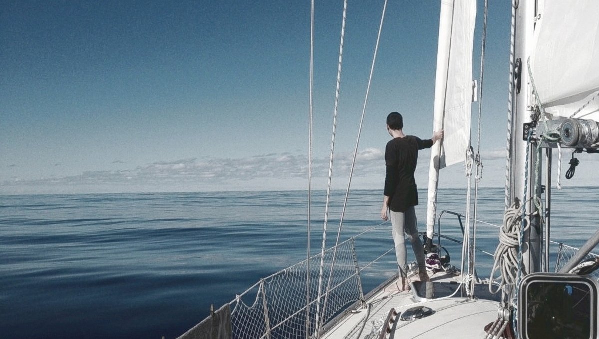 A photo of Devine standing at the bow of Pino with a quiet flat sea in the background