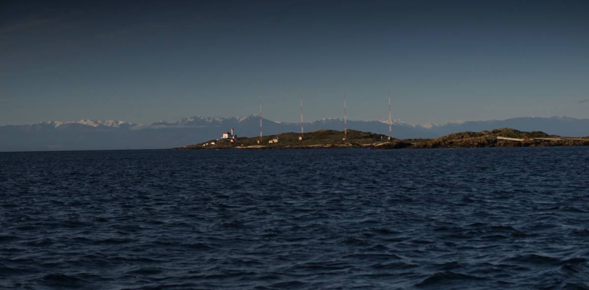 view from the deck of a sailboat of a lighthouse with snowcapped mountains in the background