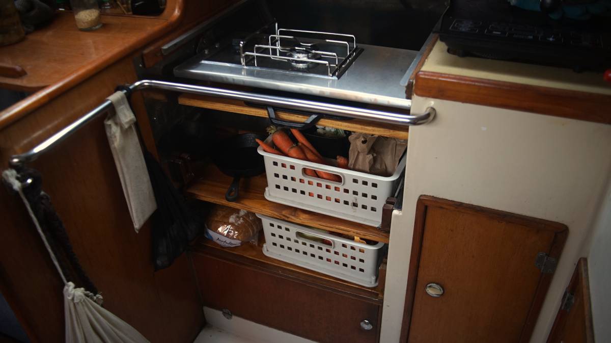 a single gimballed burner installed over two shelves containing baskets of fresh produce in a sailboat galley