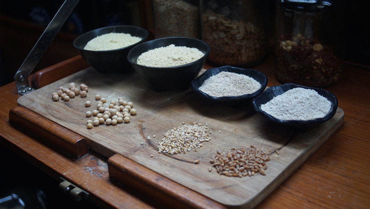 A photo featuring 4 whole grains: chickpeas, soybeans, oat groats and wheat berries in front of four bowl containing the flour versions of those same grains