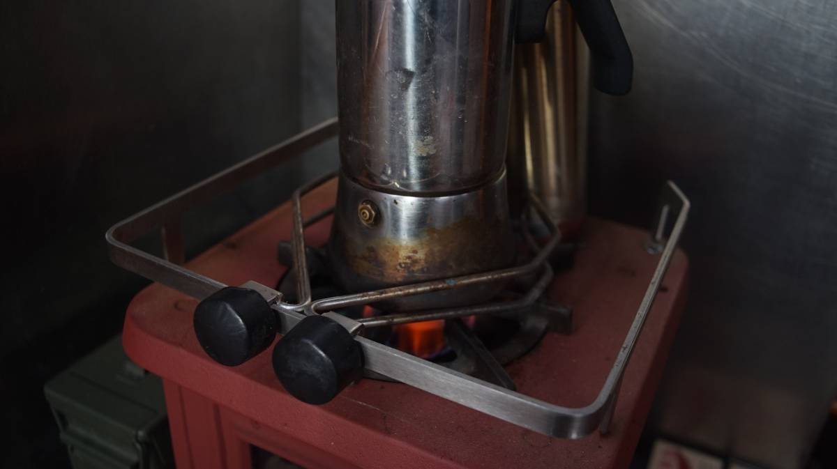 a coffee pot atop a woodstove, heated by alcohol, held by two pot holders