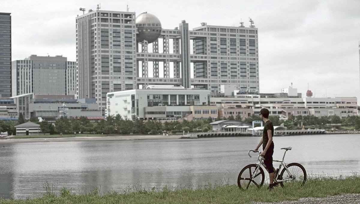 Devine is on a bike on a grassy shore, facing back to us in front of water with an imposing building with a spherical wing in the background.