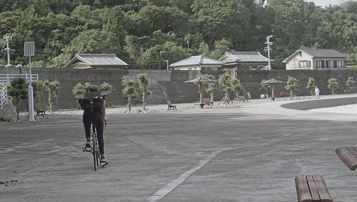 Rek is cycling over concrete walkway, a skateboard strapped to their backpack, in a small Japanese ocean-side village