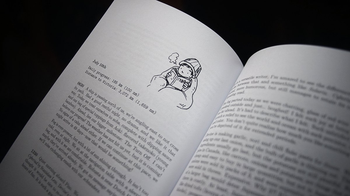 A paperback book is opened to a page featuring an illustration of Devine reading while wrapped in blankets