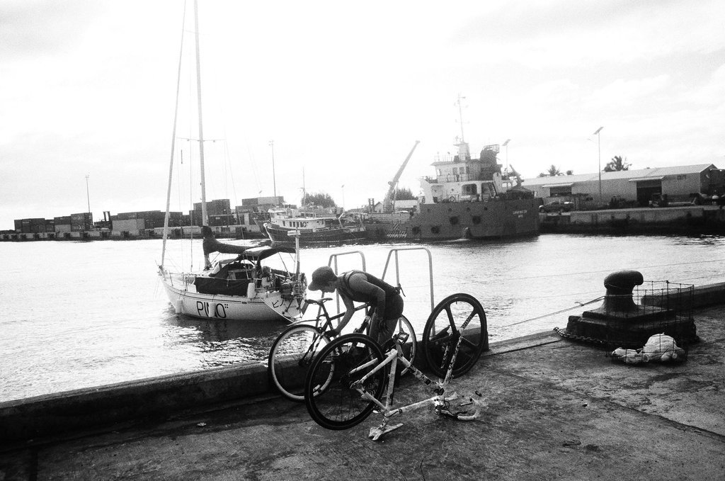 A black and white photograph of someone putting two bikes together on a concrete pier overlooking a harbour with a sailboat med-moored in the background