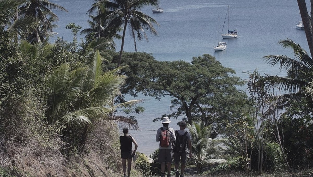A photo of 3 people walking down a step hill in Fiji, the water is in the background with a few sailboats at anchor
