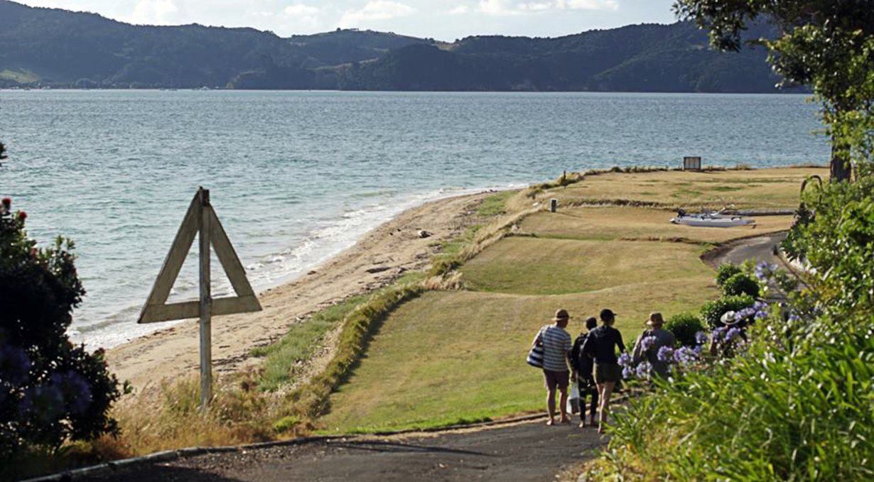 a group of people walking down a green hill, following a road with a picturesque view of the water and island in the background