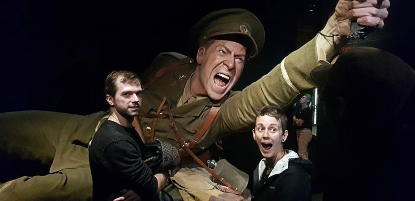 two people standing next to a giant-sized statue of a soldier in uniform, aiming a gun and screaming